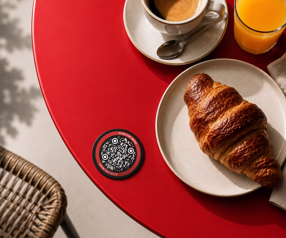 A QR code coaster on a cafe table beside an espresso and a croissant
